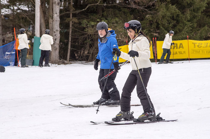 Student in white winter coat on skis and skiing across snow while a trainer in a blue winter jacket looks on.