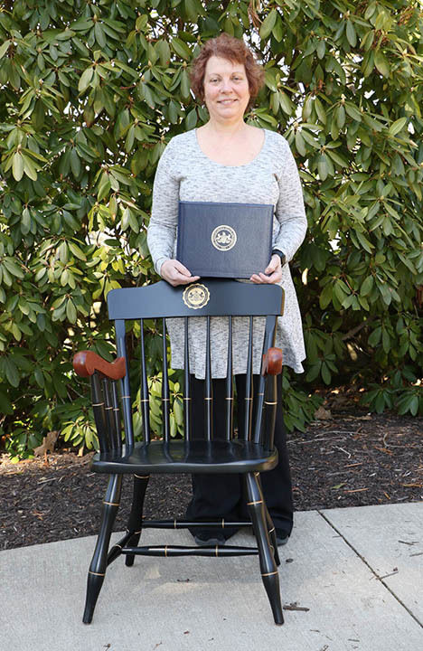 Woman standing behind chair and posing with certificate in front of green trees.