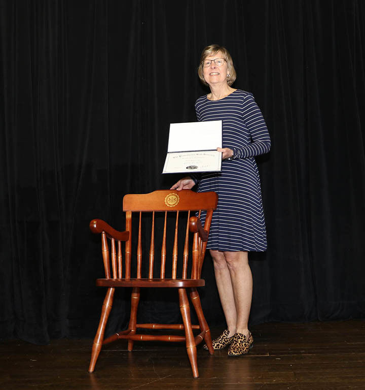 Woman holding certificate and standing in front of chair in front of a black curtain.