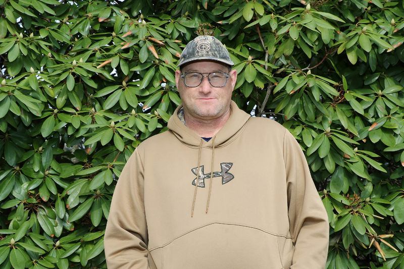 Man in hat and hooded sweatshirt standing in front of green trees.