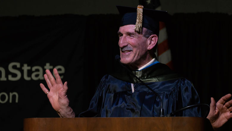 A male professor in cap and gown gestures while delivering the commencement address. 