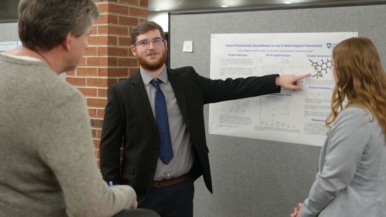 Student in a suit gesturing to a white poster board as others look on.