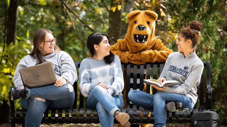 Three female students on bench with Nittany Lion mascot standing behind.