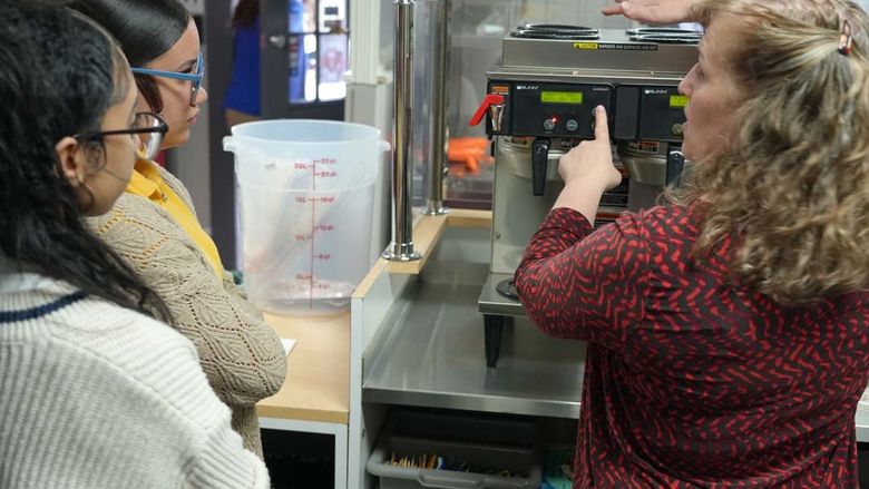 Woman holding coffee pot next to coffee machine while students observe.