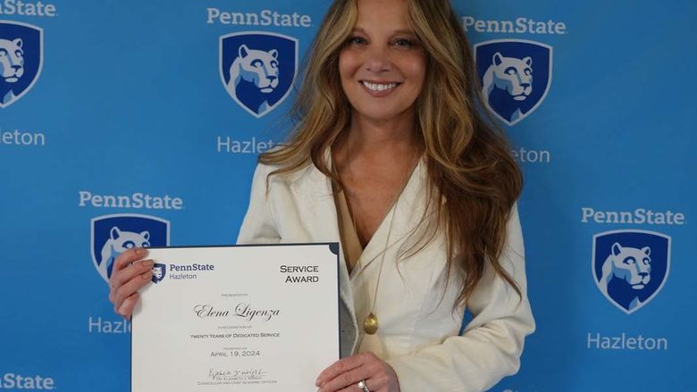 Woman with blonde hair and white jacket standing with certificate in front of light blue Penn State Hazleton backdrop.