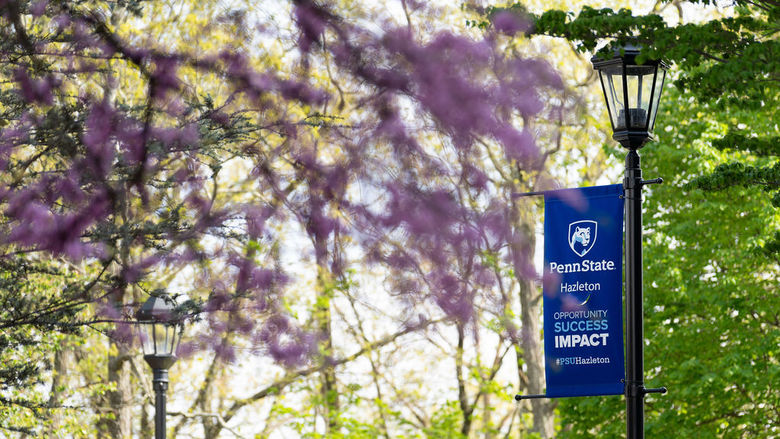A blue Penn State Hazleton banner on a lightpost with a pink flowered tree in the foreground.
