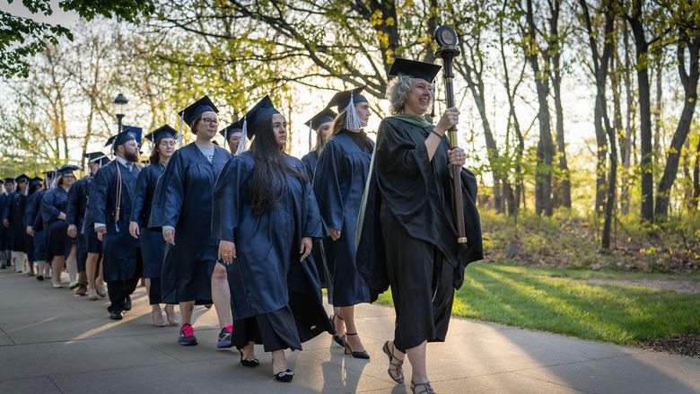 Graduates in caps and gowns being led down a tree-lined sidewalk during a processional.