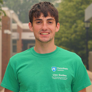 Student in green t-shirt standing outdoors in front of brick building.