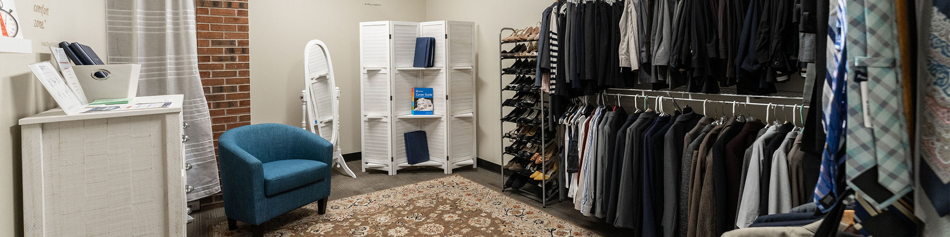 Rack of clothing including suits and ties in a room with a blue chair and white cabinets.