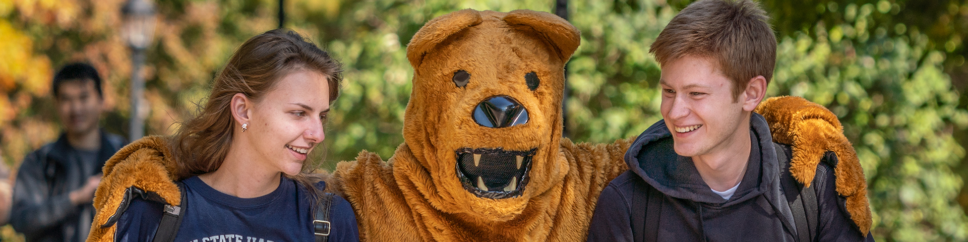 Nittany Lion mascot with arms around male and female student as the three walk together outdoors.