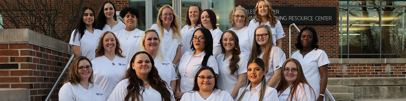 Nursing students in scrubs standing in three rows on steps outside brick building.