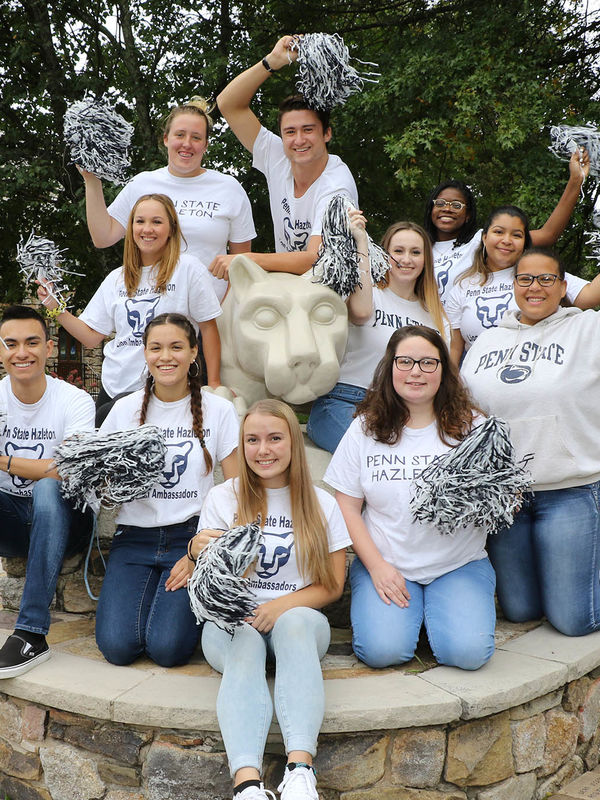 Students in white T-shirts gathered around Nittany Lion statue waving pom-poms.