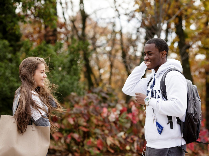 Male and female students standing across from one another in front of fall foliage.