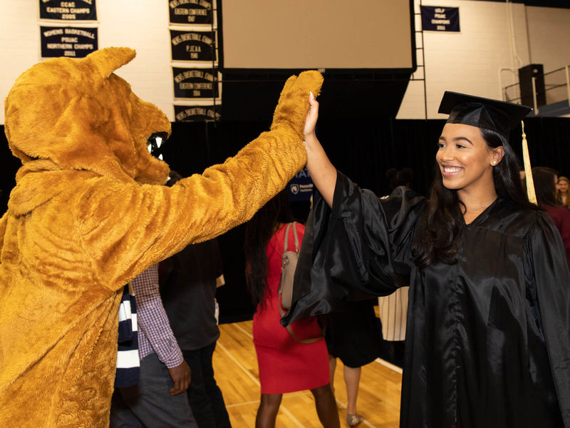 Nittany Lion mascot and female student in cap and gown high-fiving.
