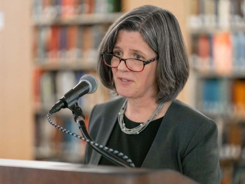 A woman in glasses smiles from behind a microphone at a podium,