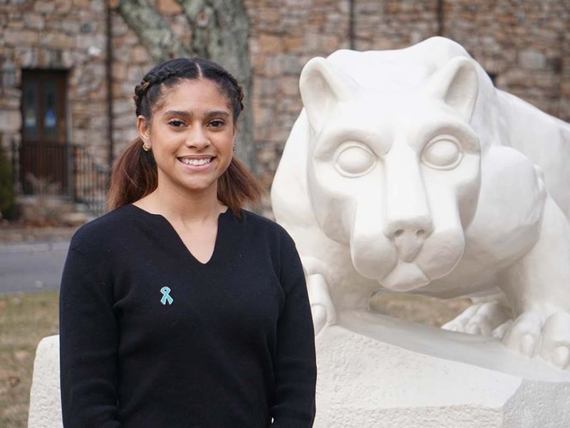 Student with dark hair in a black blouse standing in front of white Nittany Lion statue.