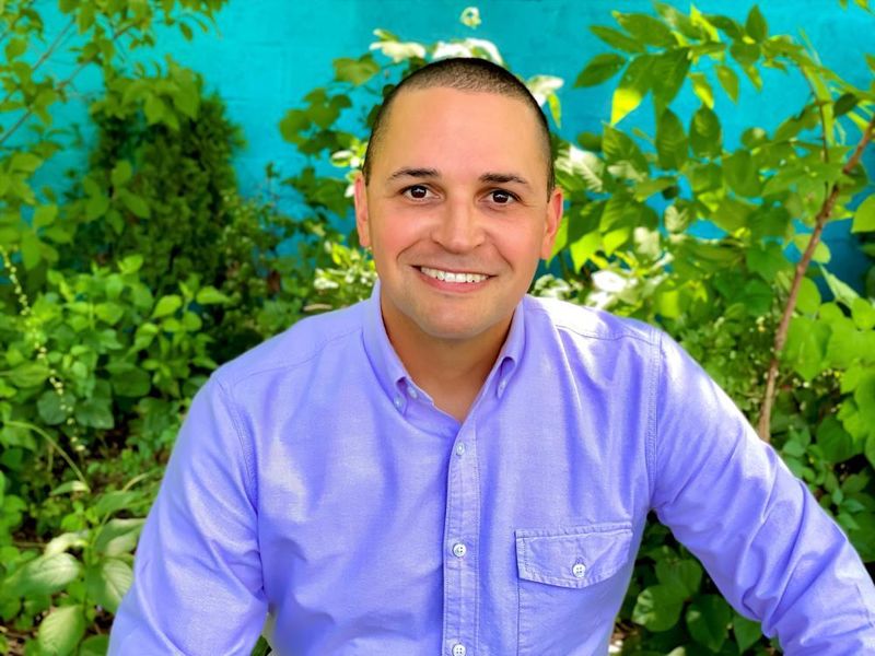 Man in a purple dress shirt seated in front of bushes and trees.