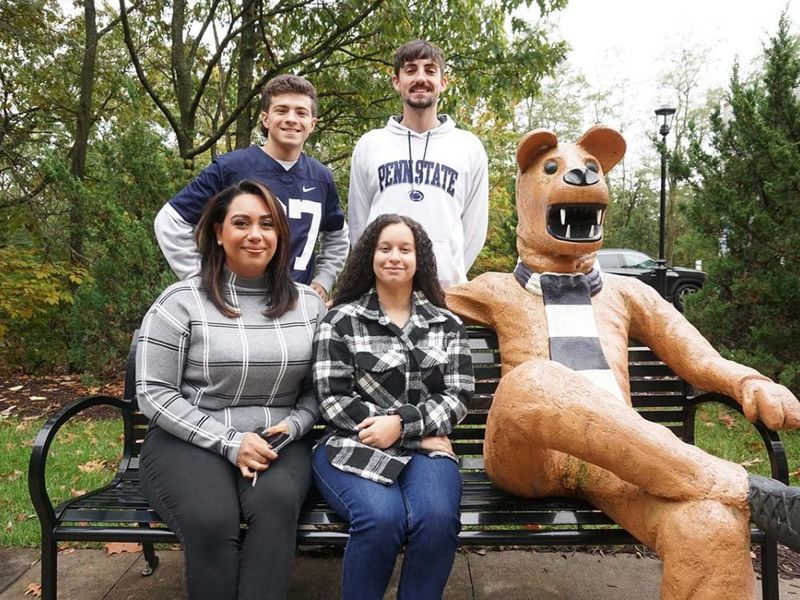 Four students gathered around a bench on a sidewalk seated next to a Nittany Lion statue.