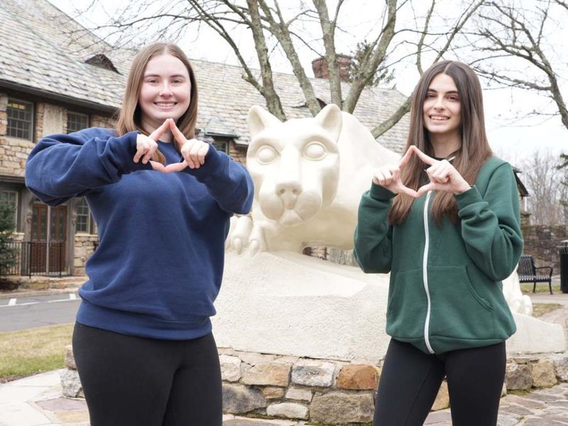 Two female students making diamond shape with their hands in front of a Nittany Lion statue.