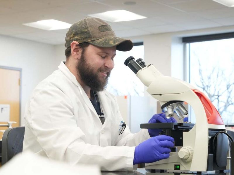 Male student in white lab coat and camouflaged baseball cap looking through a microscope in a lab 