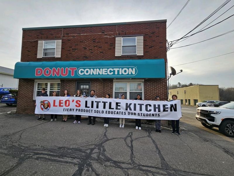Group of people holding a banner that reads "Leo's Little Kitchen" outside a storefront.