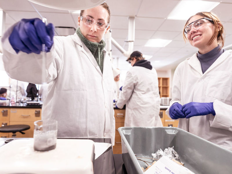 Two students in white lab coats in a chemistry laboratory mixing chemicals in a glass beaker.