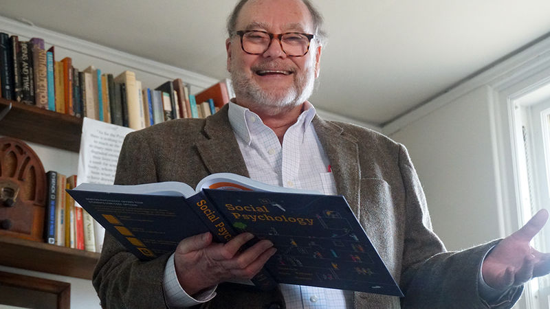 Man holding open text book standing in front of book shelf.