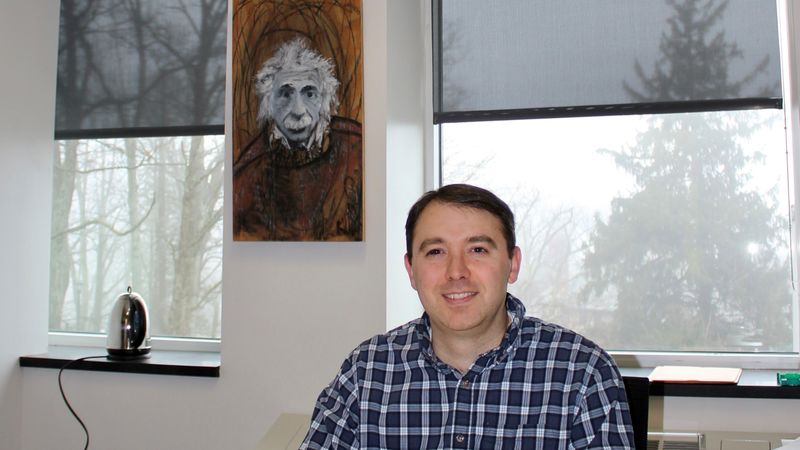 Joe Ranalli in collared shirt sitting at office desk with Albert Einstein poster in background