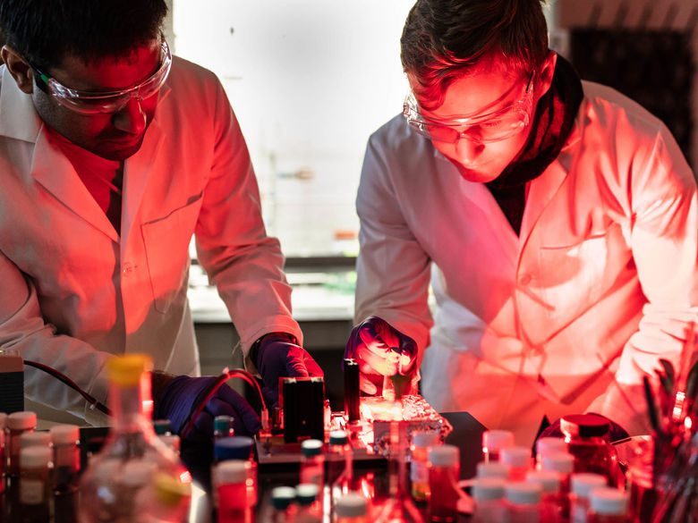Professor and student in white lab coats standing over chemistry equipment that is radiating red light.