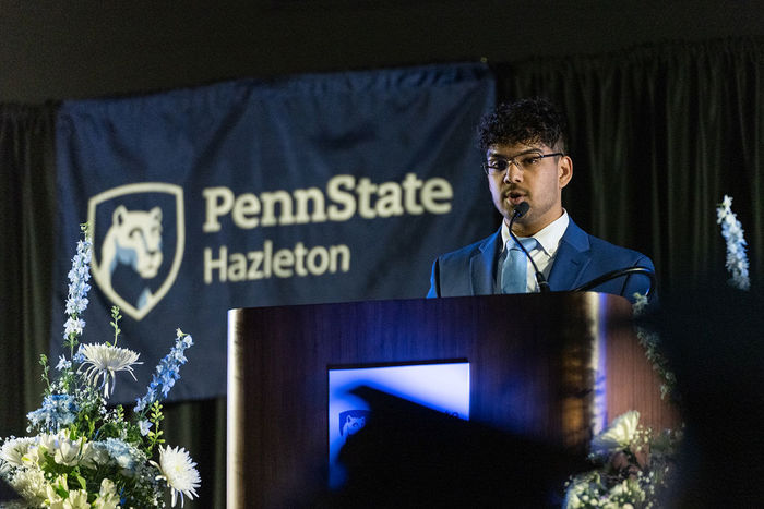Male student in a suit and tie speaking from behind a podium during a college graduation ceremony.
