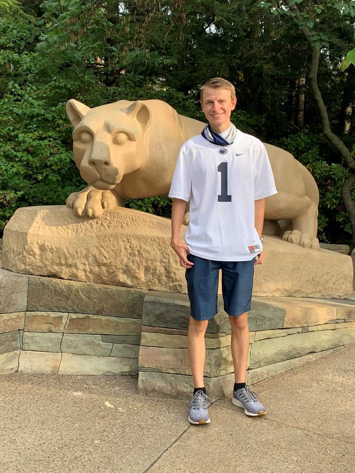 Student in Penn State football jersey standing in front of Nittany Lion statue