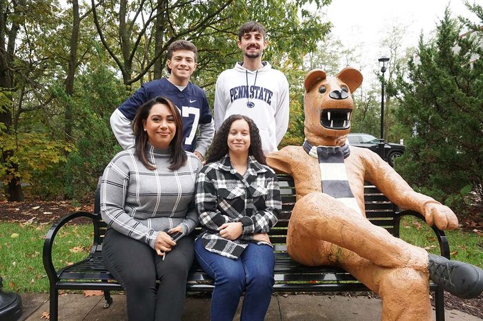 Students gathered around a bench next to the Nittany Lion.Two female students are seated with two male students standing behind them.