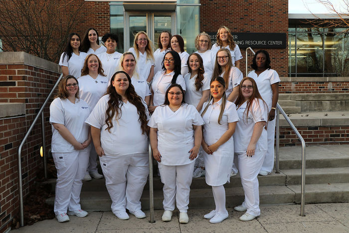 Women in nursing scrubs standing in three rows in front of brick building.