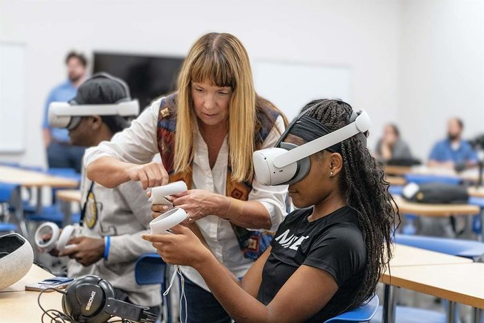 Female student wearing white virtual reality headset with female professor standing next to her offering guidance,