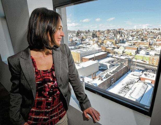Woman in gray blazer and red shirt looking over her shoulder outside a window overlooking city landscape.