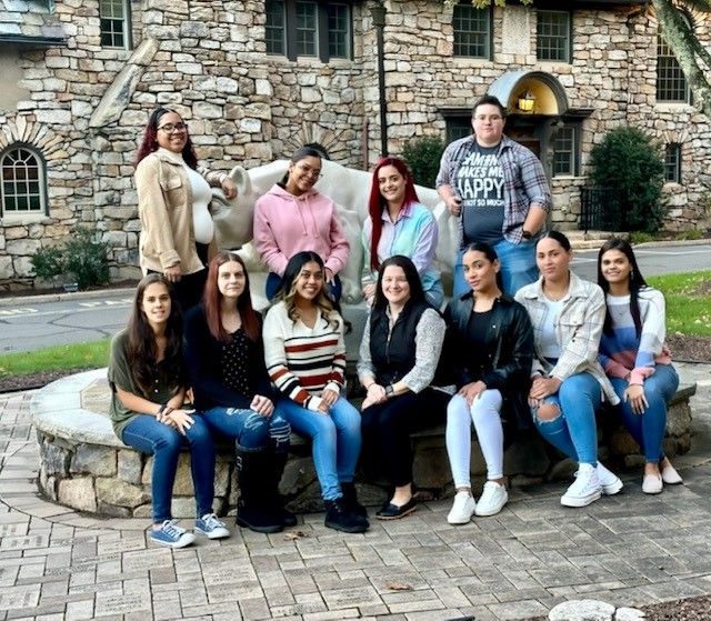 Group of female students in two rows, one row seated and the other standing, all gathered outdoors around statue of Nittany Lion.