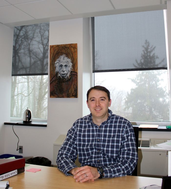 Joe Ranalli in collared shirt sitting at office desk with Albert Einstein poster in background