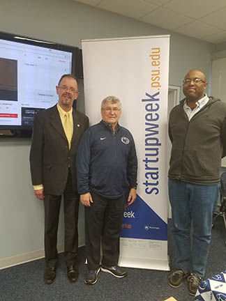 Three men standing in front of TV screen and banner. 