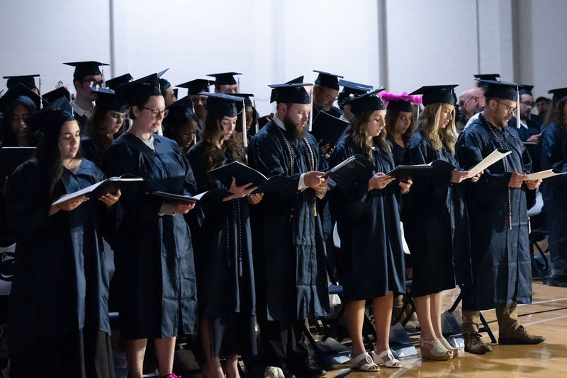 Line of graduates reading from a booklet while standing.