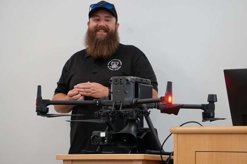 Man in baseball cap and beard standing in front of large, black drone.