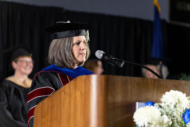 A woman in a dark colored academic regalia delivers remarks at a brown podium.