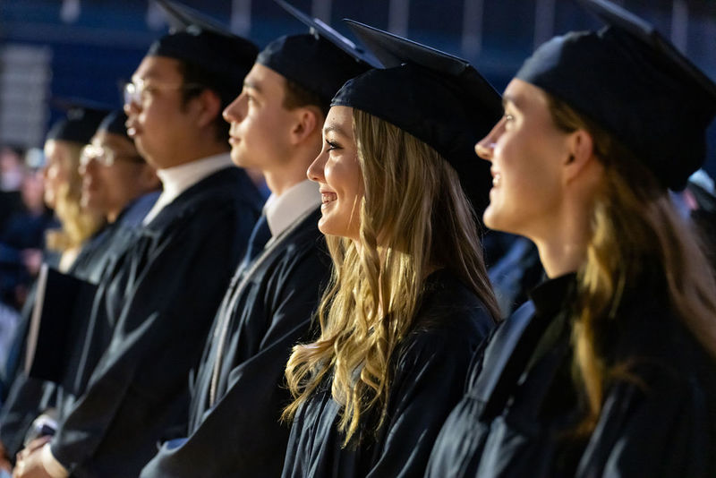 A female graduate smiles and looks toward a stage surrounded in seats by other graduates.