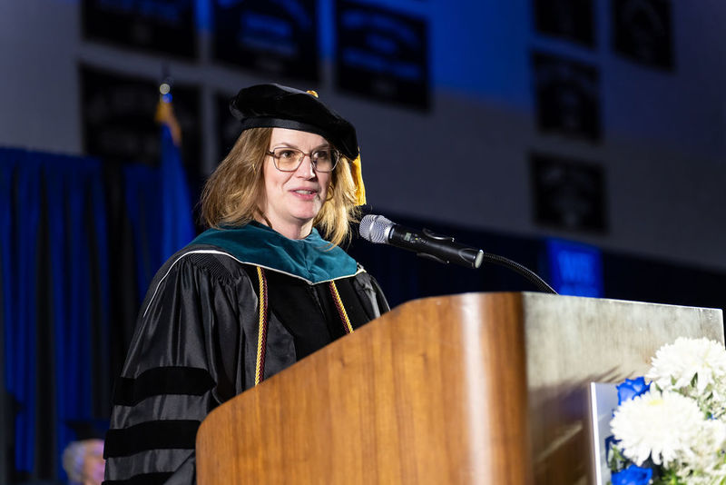 Woman with brown hair in dark academic regalia speaking at a podium.