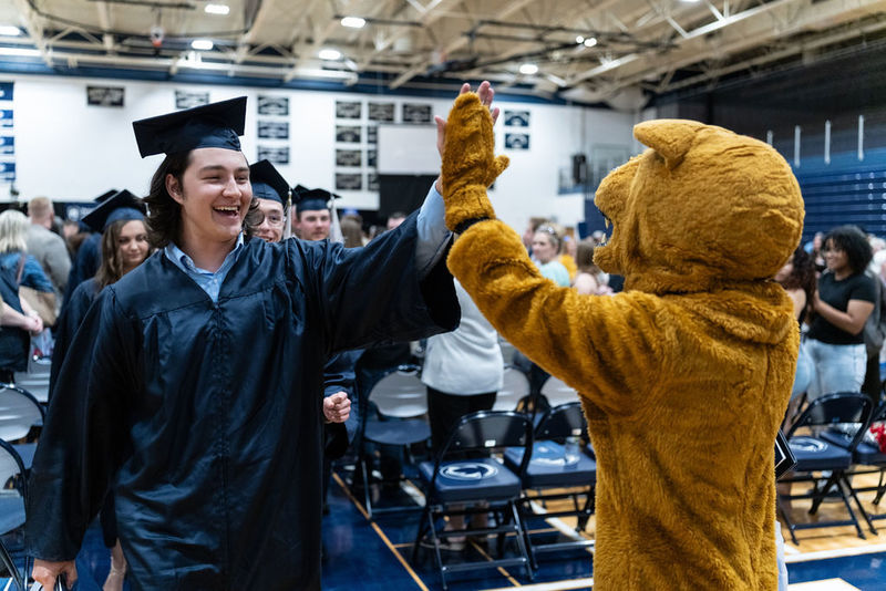 A graduate in a blue cap and gown high fives a Nittany Lion mascot.