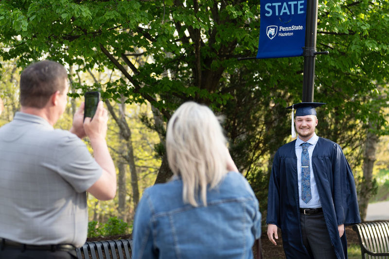 Student in cap and gown having photo taken by man and woman in front of a tree and light post.