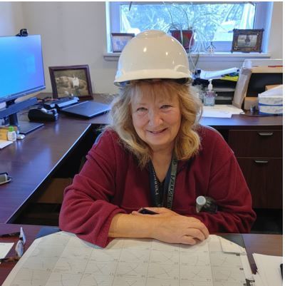 Woman sitting at a desk smiling wearing a hard hat.