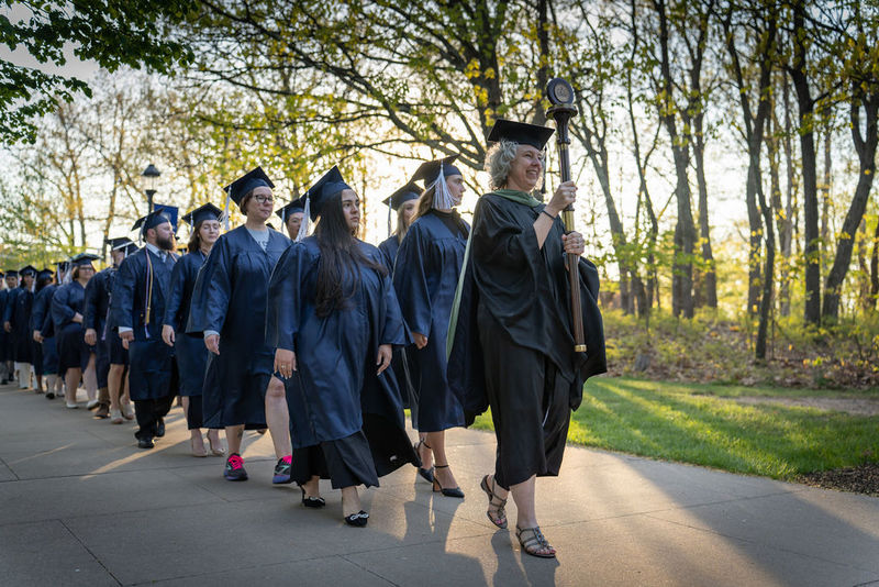 Graduates in caps and gowns being led down a tree-lined sidewalk during a processional.