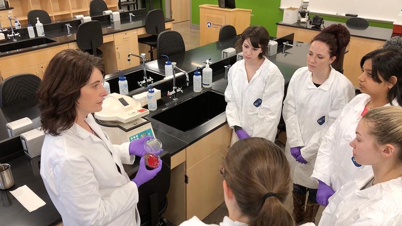 A group of students in lab coats look at a sample being held by an instructor. 