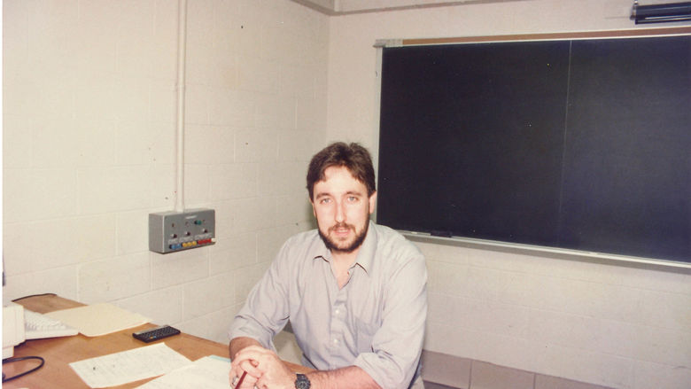 Instructor sitting at desk in classroom with chalkboard behind him.
