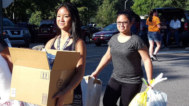 Two female students carrying boxes and bags in a parking lot.